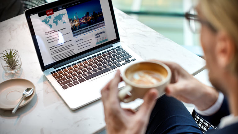 Homme avec un café en main lisant un article sur son laptop.