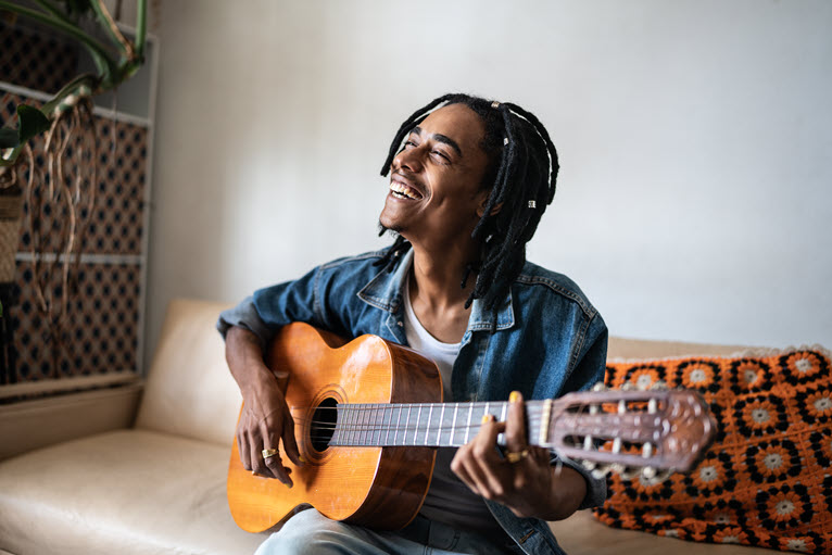 Un homme noir avec des locs dans les cheveux, souriant, assis sur un divan beige avec une guitare à la main.